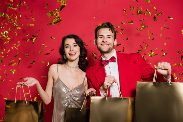 Stylish couple holding shopping bags under falling confetti on red background - Foto, afbeelding
