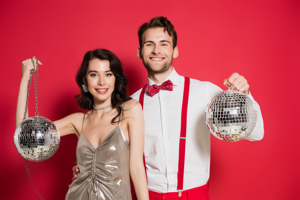 Smiling man embracing girlfriend while holding disco balls on red background - Foto, imagen