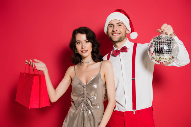 Kırmızı arka planda disko topu olan Noel Baba şapkalı sevgilisinin yanında alışveriş torbaları tutan gülümseyen bir kadın. - Fotoğraf, Görsel
