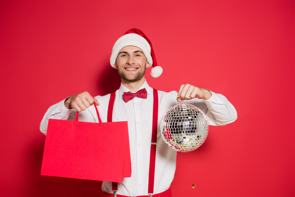 Χαμογελώντας άνθρωπος στο καπέλο Σάντα κρατώντας ντίσκο μπάλα και τσάντες ψώνια σε κόκκινο φόντο - Φωτογραφία, εικόνα