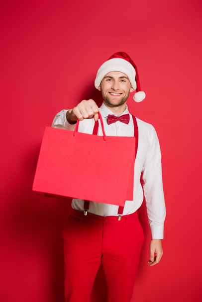 Stylish man in santa hat smiling while holding shopping bags on red background - Foto, Imagem