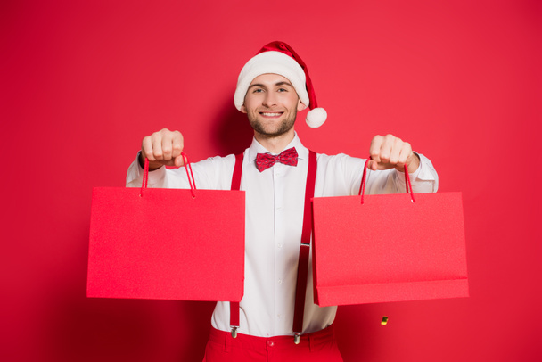 Young man in santa hat holding shopping bags and smiling at camera on red background - Fotó, kép