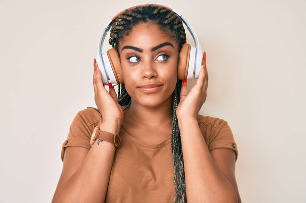 Młoda Afroamerykanka z warkoczami słuchająca muzyki za pomocą słuchawek uśmiechnięta patrząca w bok i patrząca myśląc.  - Zdjęcie, obraz