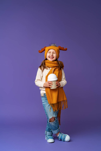 happy little girl in yellow hat, scarf with moose horns on her head stands on purple background. child smiles with his teeth, closes his eyes, holding glass with cocktail tube in form of ice cream. - Fotografie, Obrázek