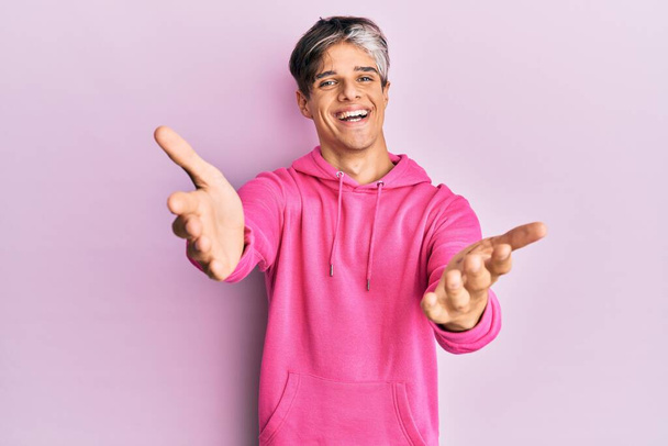Junger hispanischer Mann in lässigem rosafarbenem Sweatshirt, der in die Kamera lächelt und mit offenen Armen umarmt wird. fröhlicher Ausdruck, der das Glück umarmt.  - Foto, Bild