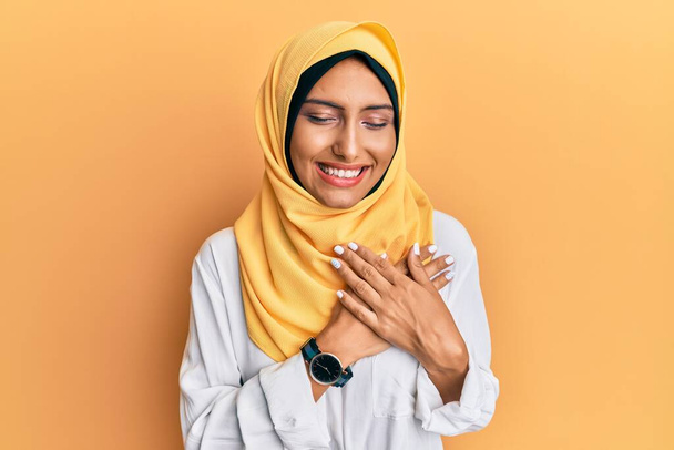 Молода брюнетка арабська жінка в традиційному ісламському шарфі хіджаб посміхається руками на грудях із закритими очима і вдячним жестом на обличчі. концепція здоров'я
.  - Фото, зображення