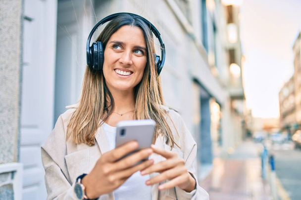 Jovem caucasiana sorrindo feliz usando smartphone e fones de ouvido na cidade. - Foto, Imagem
