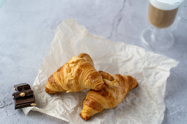 On a gray table there is a glass cup with hot latte coffee and next to it.Two croissants lie on a gray background next to three pieces of chocolate and black candles - Photo, image
