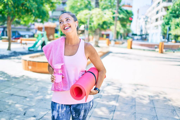 Middelbare leeftijd sportvrouw glimlachend gelukkig houden yoga mat en fles water in het park - Foto, afbeelding