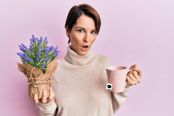 Young brunette woman with short hair drinking a cup of infused lavender in shock face, looking skeptical and sarcastic, surprised with open mouth  - Photo, Image