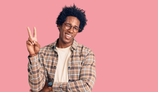 Jóképű afro-amerikai férfi afro hajjal, alkalmi ruhában és szemüvegben, mosolyogva, boldog arccal kacsintva a kamerára, győzelmi jelet téve az ujjaival. Második..  - Fotó, kép