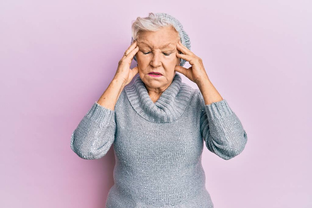 Eine ältere grauhaarige Frau mit Wollpullover und Wintermütze leidet an Kopfschmerzen und ist gestresst, weil sie Schmerzen und Migräne hat. Hände auf den Kopf.  - Foto, Bild