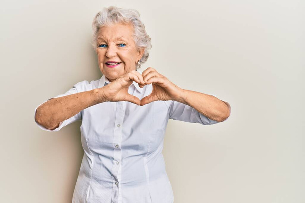 Starší šedovlasá žena v neformálním oblečení, usmívající se v lásce, dělá rukou tvar symbolu srdce. romantický koncept.  - Fotografie, Obrázek