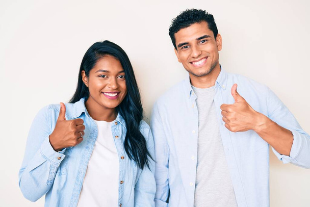 Beautiful latin young couple wearing casual clothes success sign doing positive gesture with hand, thumbs up smiling and happy. cheerful expression and winner gesture.  - Fotografie, Obrázek