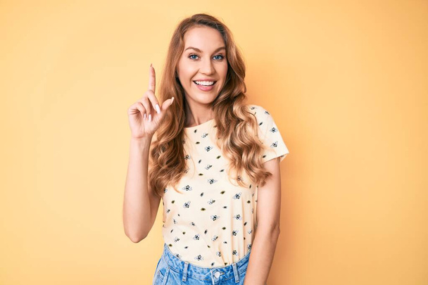 Mulher caucasiana jovem com cabelo loiro vestindo roupas de verão casuais sorrindo com uma ideia ou pergunta apontando o dedo para cima com o rosto feliz, número um  - Foto, Imagem