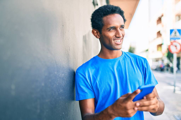 Όμορφος μαύρος Αφρικανός άντρας χαμογελά χαρούμενος σε εξωτερικούς χώρους χρησιμοποιώντας το smartphone - Φωτογραφία, εικόνα