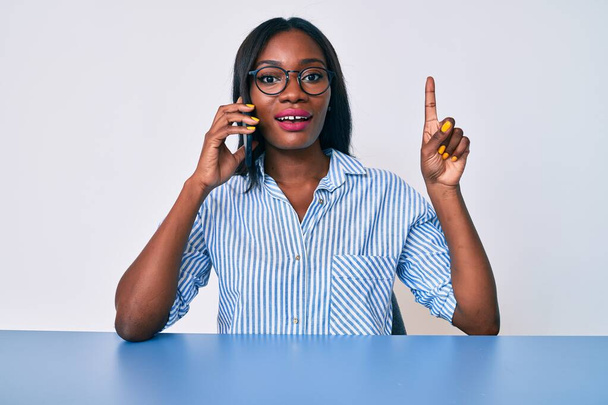 Молодая африканская американка, говорящая на смартфоне, сидя на столе, удивлена идеей или вопросом, указывая пальцем со счастливым лицом, номер один  - Фото, изображение