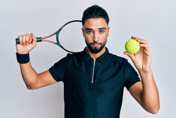 Молодой человек с бородой играет в теннис держа ракетку и мяч расслабленным с серьезным выражением лица. простой и естественный взгляд на камеру.  - Фото, изображение