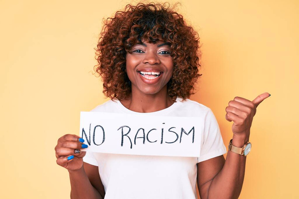Joven mujer afroamericana sin pancarta de racismo apuntando con el pulgar hacia un lado sonriendo feliz con la boca abierta  - Foto, imagen
