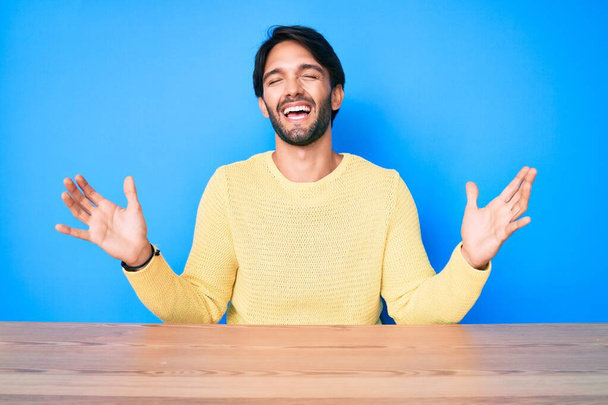 Jóképű spanyol férfi alkalmi pulóvert visel az asztalon, őrülten és őrülten ünnepli a sikert, felemelt karokkal és izgatottan sikoltozó csukott szemmel. győztes koncepció  - Fotó, kép