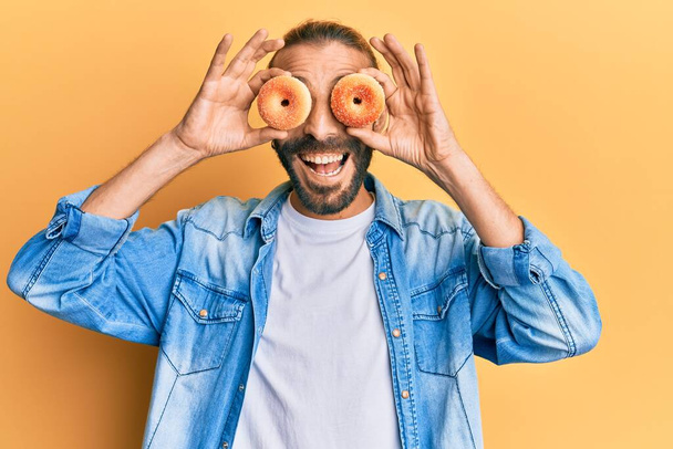 Homem atraente com cabelos longos e barba segurando saborosos donuts coloridos nos olhos sorrindo com um sorriso feliz e legal no rosto. mostrando dentes.  - Foto, Imagem