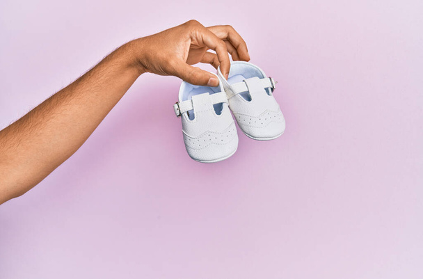Hispanic hand holding baby shoes over isolated pink background. - Photo, Image