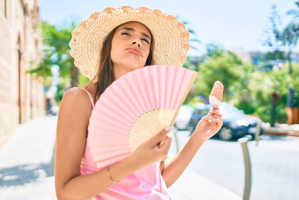 Young hispanic woman on vacation wearing summer style using hand fan at street of city - Photo, Image