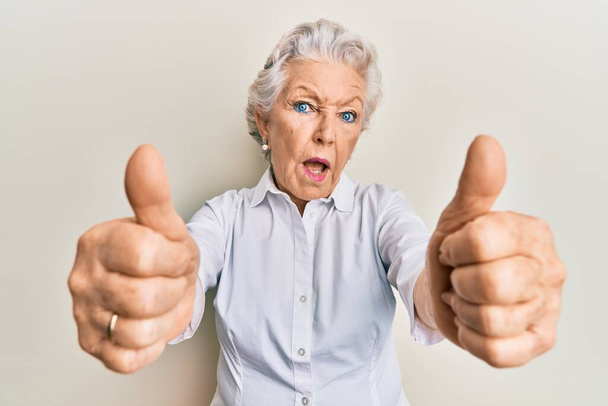 Mujer mayor de cabello gris haciendo pulgares hacia arriba gesto positivo en la cara de choque, mirando escéptico y sarcástico, sorprendido con la boca abierta  - Foto, imagen