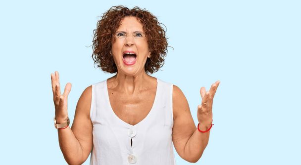 Beautiful middle age mature woman wearing casual white shirt crazy and mad shouting and yelling with aggressive expression and arms raised. frustration concept.  - Photo, Image
