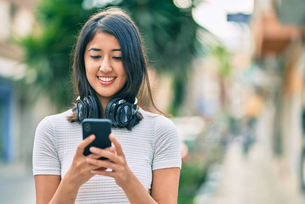 Młoda Latynoska uśmiechnięta szczęśliwa korzystając ze smartfona i słuchawek w mieście. - Zdjęcie, obraz