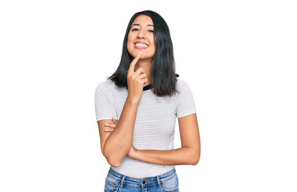 Schöne asiatische junge Frau trägt legeres weißes T-Shirt und schaut selbstbewusst in die Kamera, mit verschränkten Armen und erhobener Hand am Kinn. Positives Denken.  - Foto, Bild