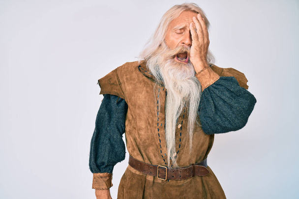 Starý starší muž s šedivými vlasy a dlouhými vousy, oblečený v tradičním vikingském kostýmu, unaveně zírající, zakrývající půlku obličeje, oko a ústa rukou. bolest v obličeji.  - Fotografie, Obrázek