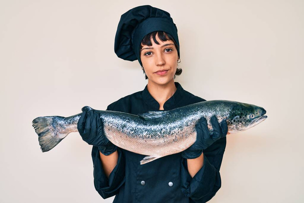 Όμορφη brunette γυναίκα επαγγελματίας σεφ κρατώντας φρέσκο ψάρι σολομό χαλαρή με σοβαρή έκφραση στο πρόσωπο. απλή και φυσική κοιτάζοντας την κάμερα.  - Φωτογραφία, εικόνα