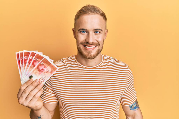 Young caucasian man holding 20 israel shekels banknotes looking positive and happy standing and smiling with a confident smile showing teeth  - Photo, image