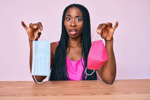 Африканская американка с косичками, держащая две разные защитные маски в шокирующем лице, выглядящая скептически и саркастично, удивленная открытым ртом  - Фото, изображение