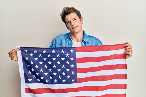 Un bell'uomo caucasico che tiene la bandiera degli Stati Uniti, espressione insensata e confusa. concetto di dubbio.  - Foto, immagini