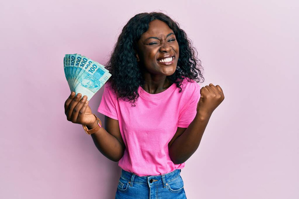 Piękna czarna kobieta trzymająca 100 brazylijskich banknotów świętujących osiągnięcie ze szczęśliwym uśmiechem i wyrazem zwycięzcy z podniesioną ręką  - Zdjęcie, obraz