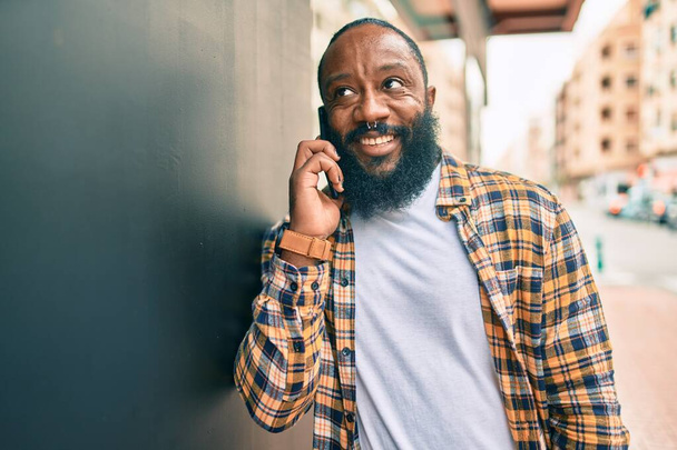 Jóképű, modern afro-amerikai férfi, szakállal a telefonban beszél a városban, boldog mosollyal. - Fotó, kép