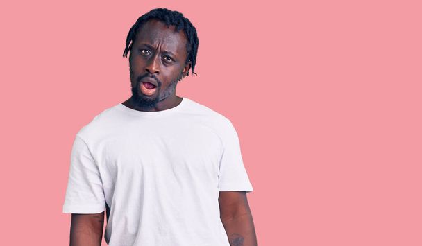 Young african american man with braids wearing casual white tshirt in shock face, looking skeptical and sarcastic, surprised with open mouth  - Fotoğraf, Görsel