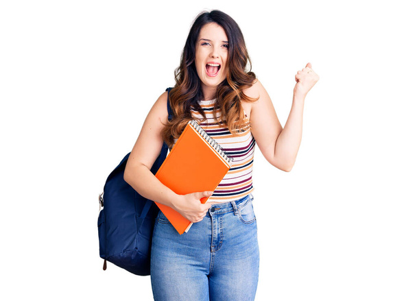 Beautiful young brunette woman wearing student backpack hodling books screaming proud, celebrating victory and success very excited with raised arms  - Foto, immagini