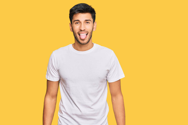 Mladý pohledný muž v nenucené bílé košili vystrkující jazyk se veselým výrazem. koncepce emocí.  - Fotografie, Obrázek