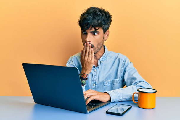 Молодой латиноамериканец, работающий в офисе, пьет чашку кофе, закрывая рот рукой, шокирован и боится ошибиться. Удивительное выражение  - Фото, изображение