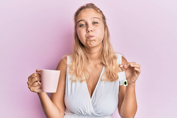 Menina loira jovem segurando xícara de chá soprando bochechas com rosto engraçado. boca inflada com ar, apanhar ar.  - Foto, Imagem