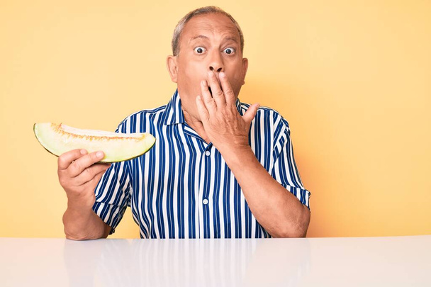 Älterer gutaussehender Mann mit grauen Haaren und frischer Melone auf dem Tisch sitzend, den Mund mit der Hand bedeckt, schockiert und ängstlich vor Irrtum. Überraschte Miene  - Foto, Bild