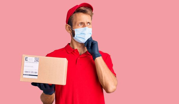 Όμορφος ξανθός άντρας με γενειάδα να κρατάει το κουτί παράδοσης φορώντας ιατρική μάσκα σοβαρό πρόσωπο σκεπτόμενος την ερώτηση με το χέρι στο πηγούνι, σκεπτόμενος την μπερδεμένη ιδέα  - Φωτογραφία, εικόνα