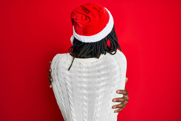 African american man with braids wearing christmas hat hugging oneself happy and positive from backwards. self love and self care  - Foto, Imagen