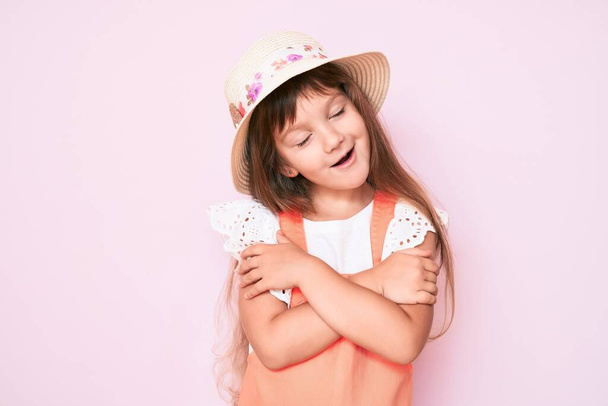 Pequeña niña caucásica con el pelo largo con sombrero de primavera abrazándose feliz y positivo, sonriendo confiado. amor propio y cuidado personal  - Foto, Imagen