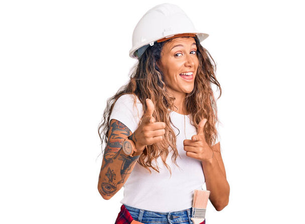 Young hispanic woman with tattoo wearing hardhat and builder clothes pointing fingers to camera with happy and funny face. good energy and vibes.  - Photo, Image