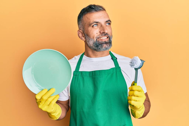 Middelbare leeftijd knappe man draagt schort met geurmaker afwas lachend kijken naar de zijkant en staren weg denken.  - Foto, afbeelding
