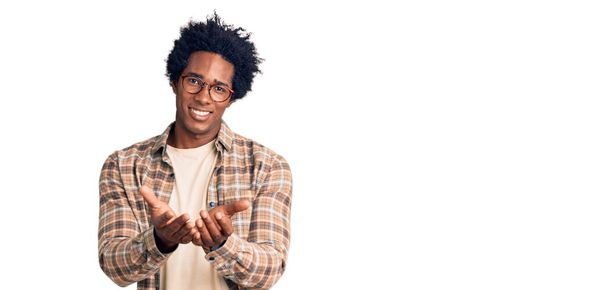 Jóképű afro-amerikai férfi afro hajjal, alkalmi ruhákat és szemüveget visel, és tenyérrel mosolyog, miközben gesztikulál. tartás és védelem  - Fotó, kép